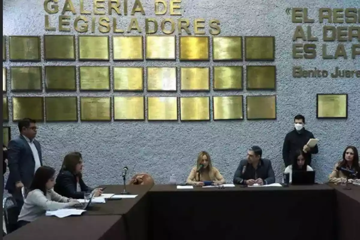 A group of people is gathered in a conference room with a background wall that has golden plaques and the words 