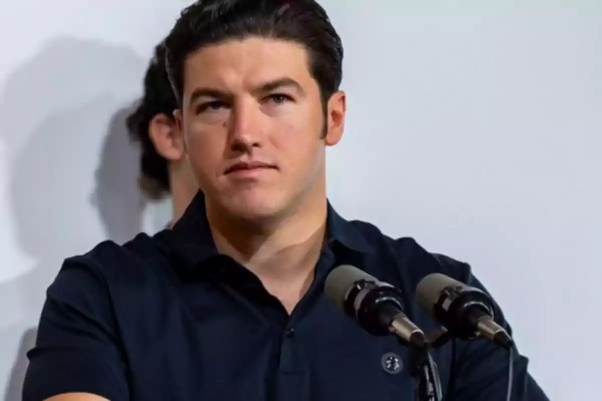 A dark-haired man in a black jersey is standing in front of a microphone.