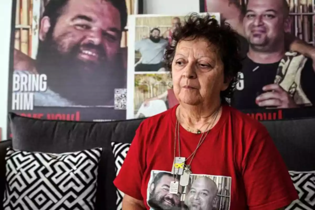 Una mujer mayor con una camiseta roja que tiene una foto impresa, sentada en un sofá con cojines de diseño geométrico, detrás de ella hay carteles con imágenes de hombres.