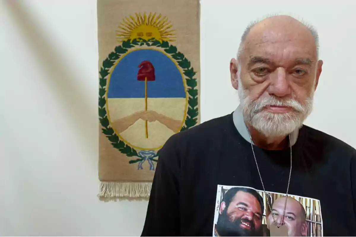 Un hombre mayor con barba blanca lleva una camiseta con una imagen de dos personas y está de pie frente a un tapiz con un escudo que tiene un sol y un gorro frigio.