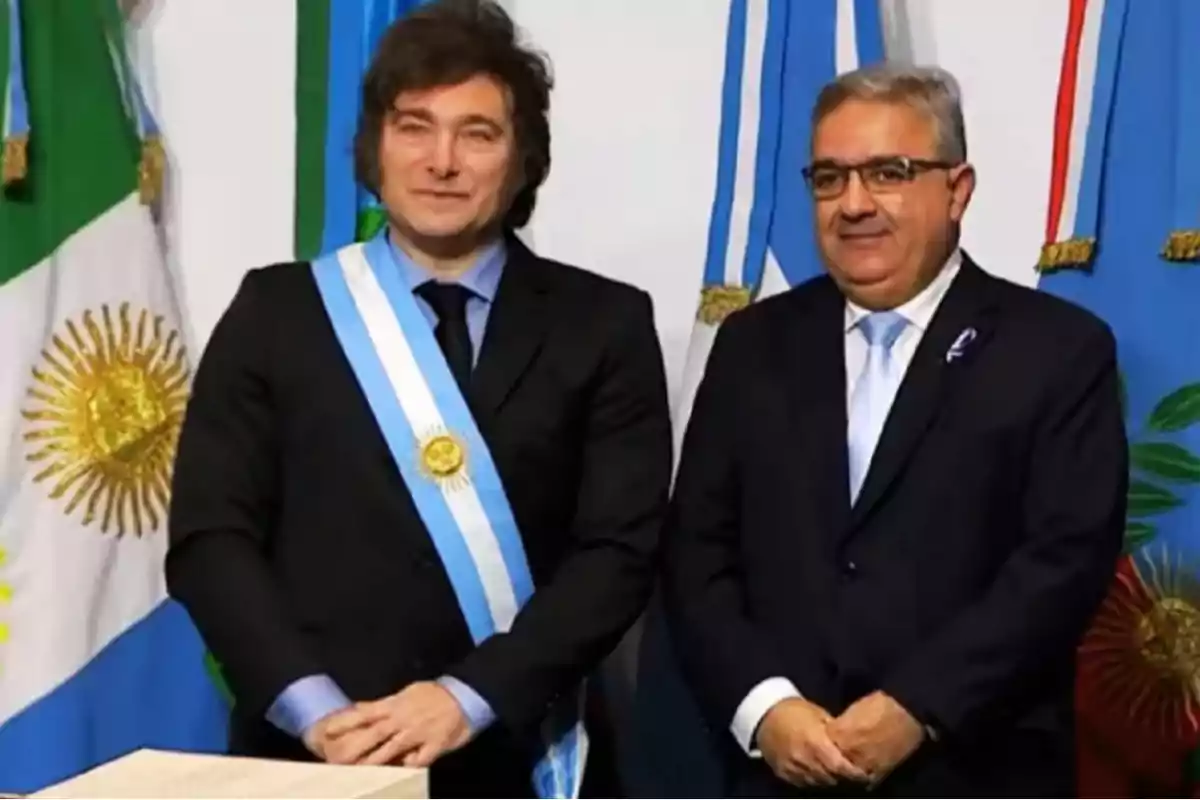 Two men standing in formal suits and presidential sashes in front of Argentine flags.