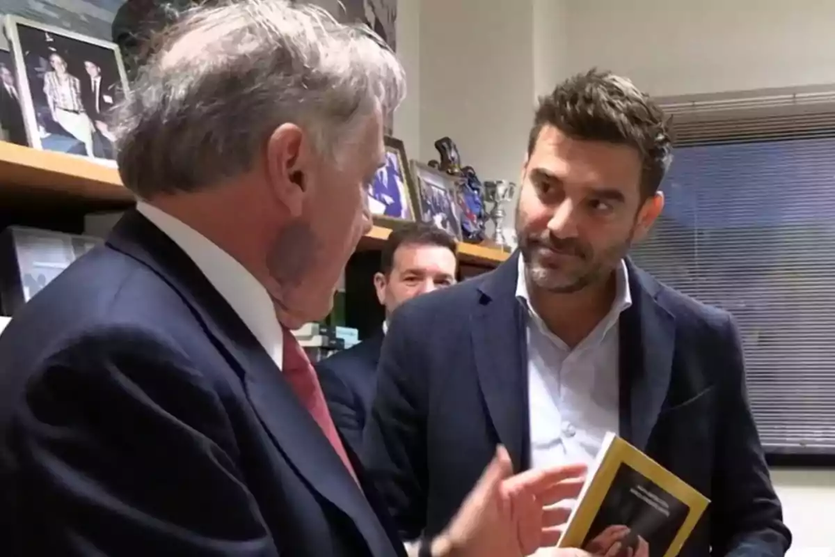 Dos hombres conversan en una oficina, uno de ellos sostiene un libro mientras el otro gesticula con la mano.