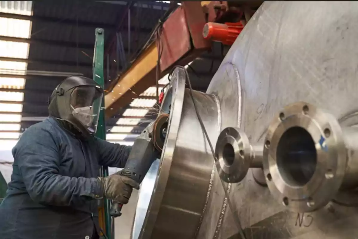 Worker with protective equipment using a power tool to work on a large metal structure in an industrial workshop.