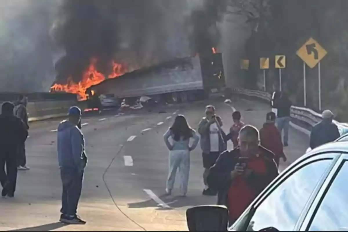 A truck on fire blocks a road while several people watch and take photos.