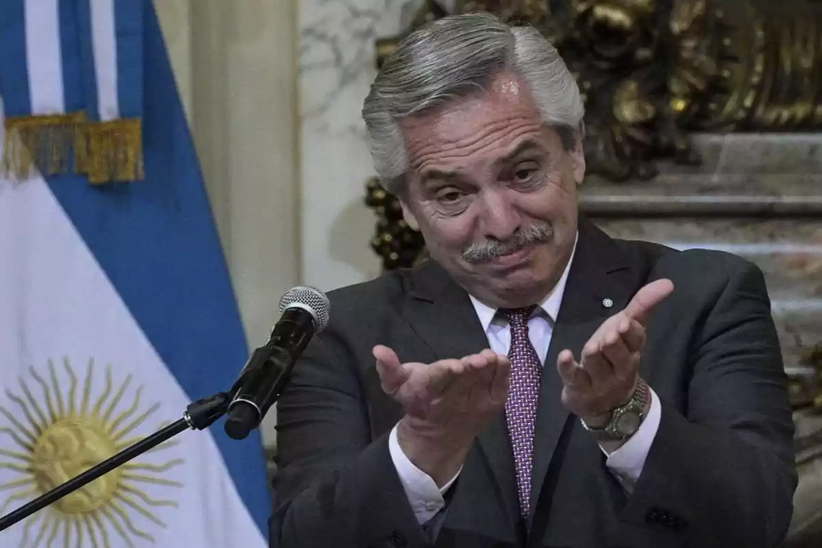 A man in a suit gestures with his hands in front of a microphone with an Argentine flag in the background.