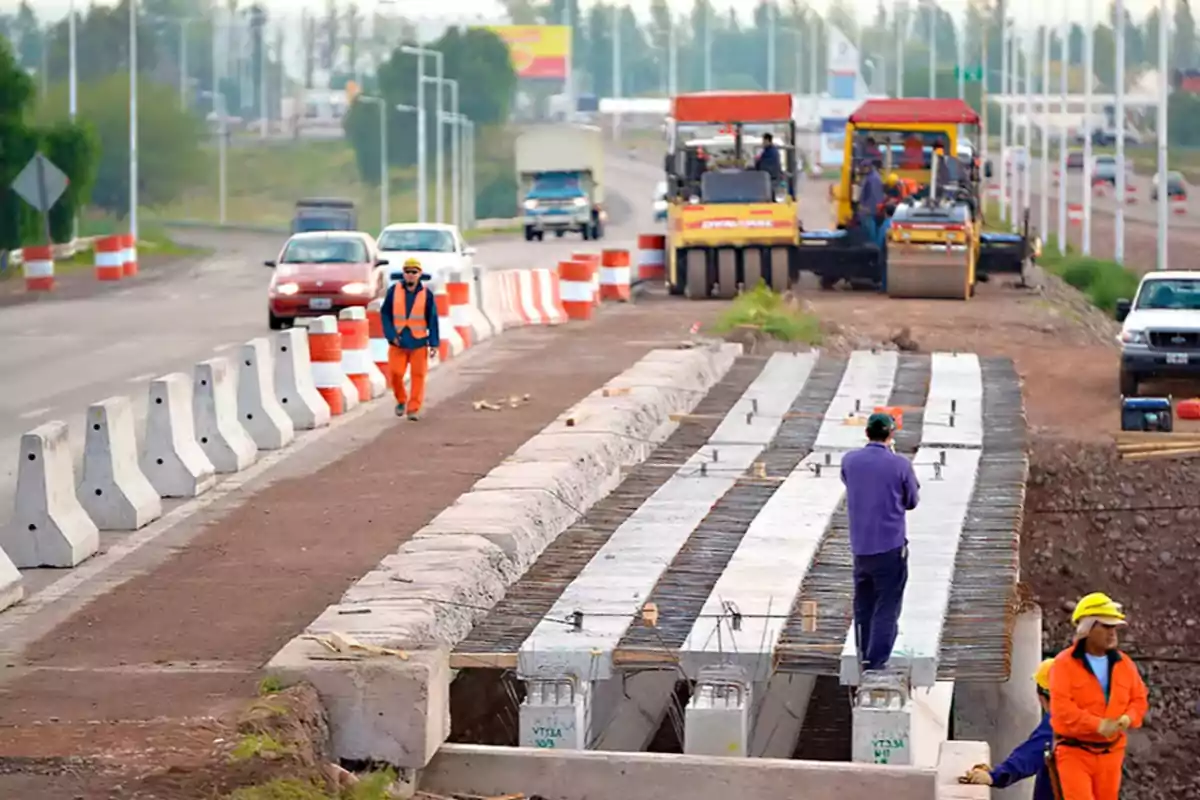 Construction workers on a road under repair, with heavy machinery and safety barriers on the sides.