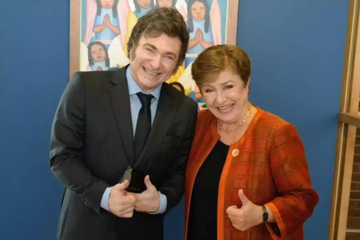 Two smiling people pose together giving a thumbs-up gesture, with a blue background and a colorful painting behind them.