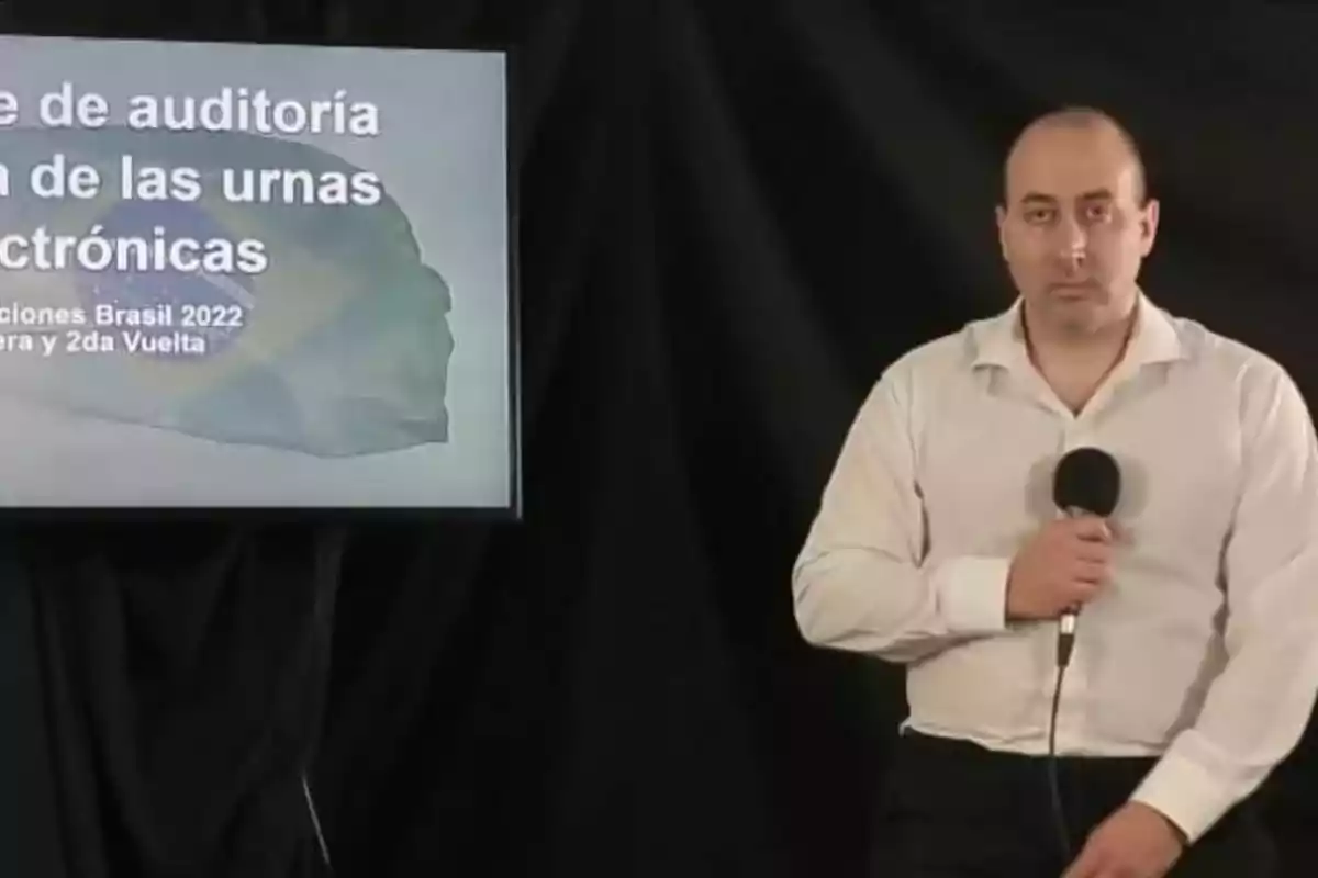 Un hombre de pie con una camisa blanca sostiene un micrófono frente a una pantalla que muestra texto relacionado con una auditoría de urnas electrónicas en las elecciones de Brasil 2022.