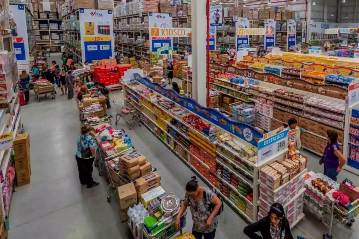 Personas comprando en un supermercado mayorista con estantes llenos de productos y carritos de compras.