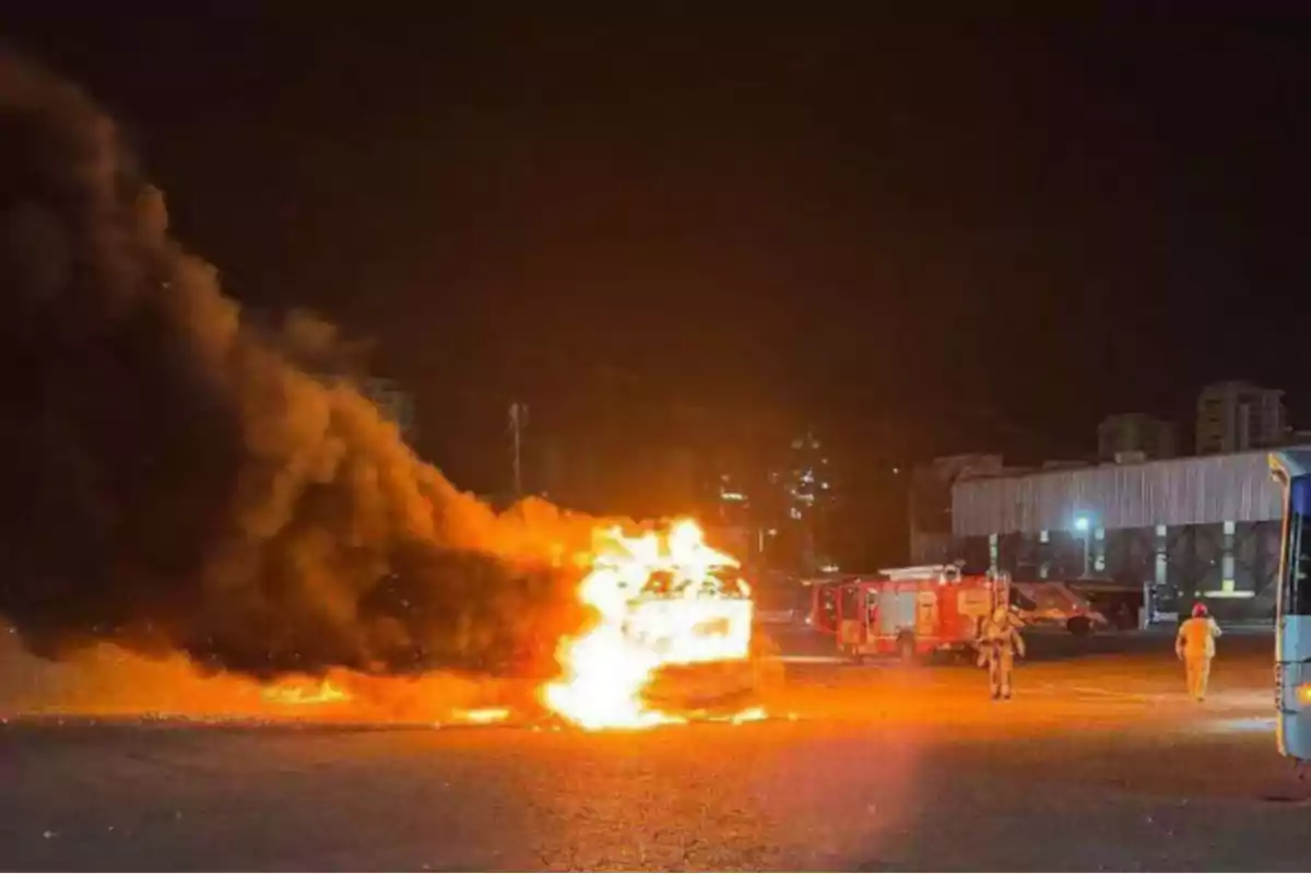 Un vehículo en llamas emite una densa columna de humo negro en un área abierta durante la noche mientras los bomberos se acercan con camiones de emergencia.