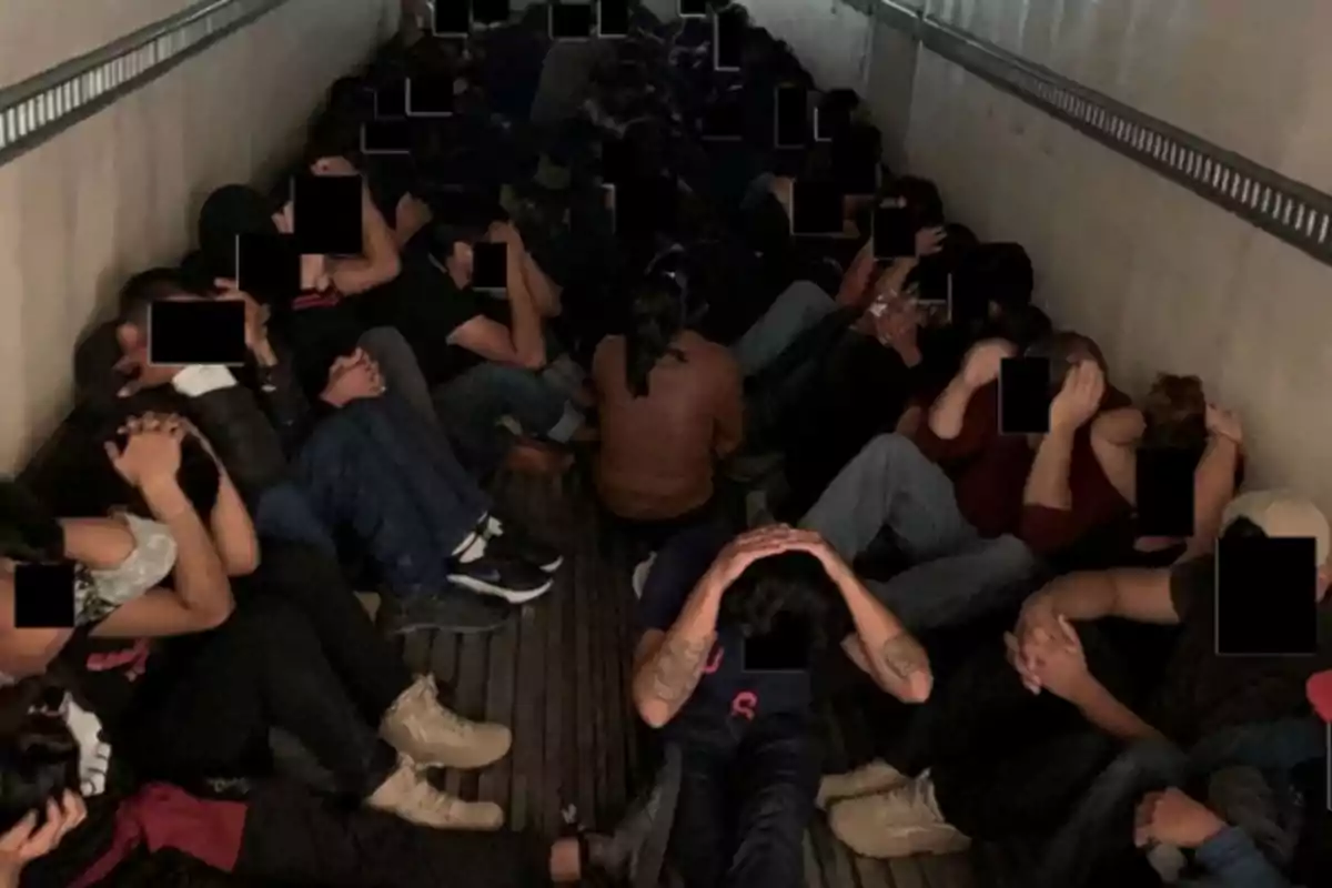 A group of people sitting on the floor of an enclosed space, with their hands on their heads and their faces pixelated.