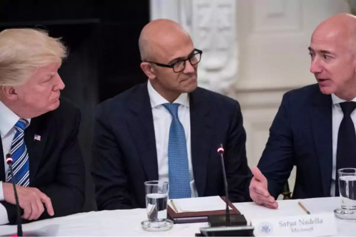 Tres hombres en trajes formales sentados en una mesa durante una reunión, con micrófonos y vasos de agua frente a ellos.