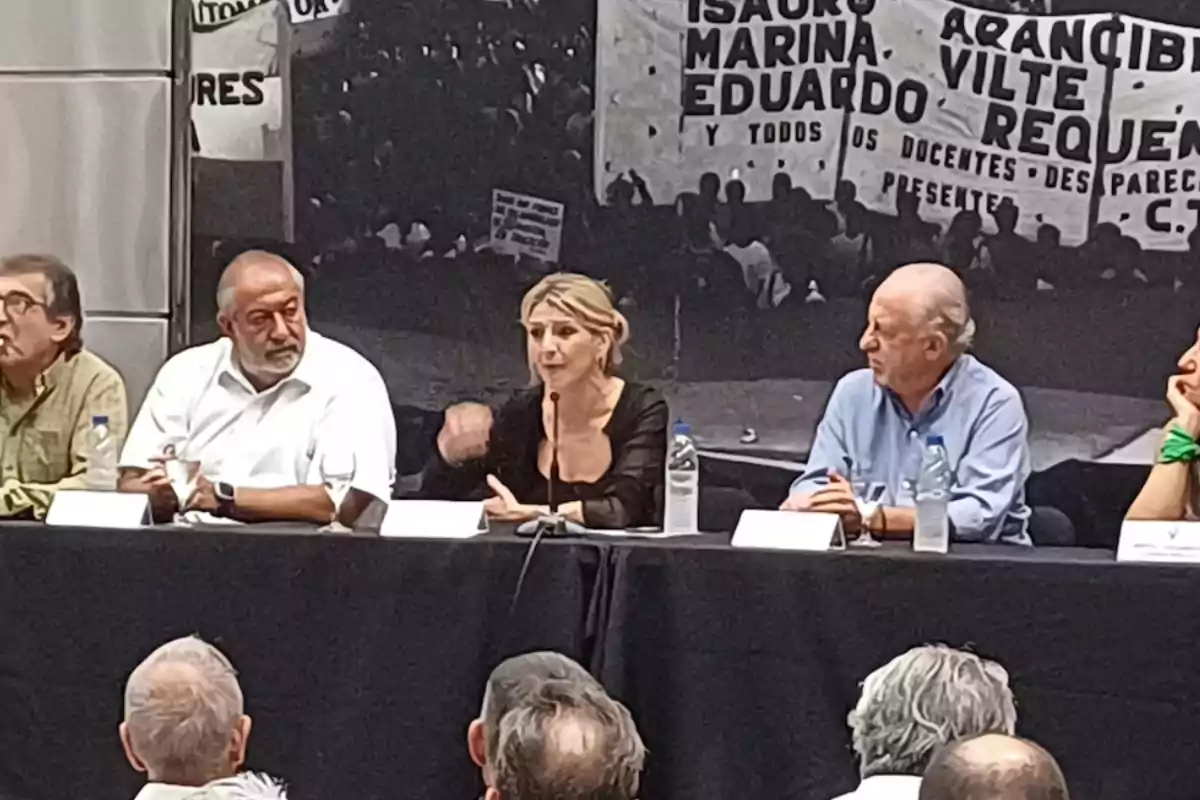 Un grupo de personas está sentado en una mesa larga durante un evento o conferencia, con un fondo que muestra una pancarta con nombres y mensajes.