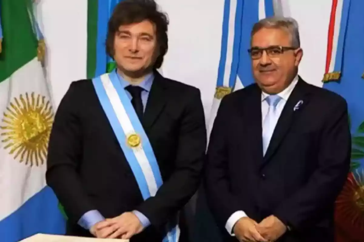 Dos hombres de pie con trajes formales y bandas presidenciales, frente a banderas argentinas.