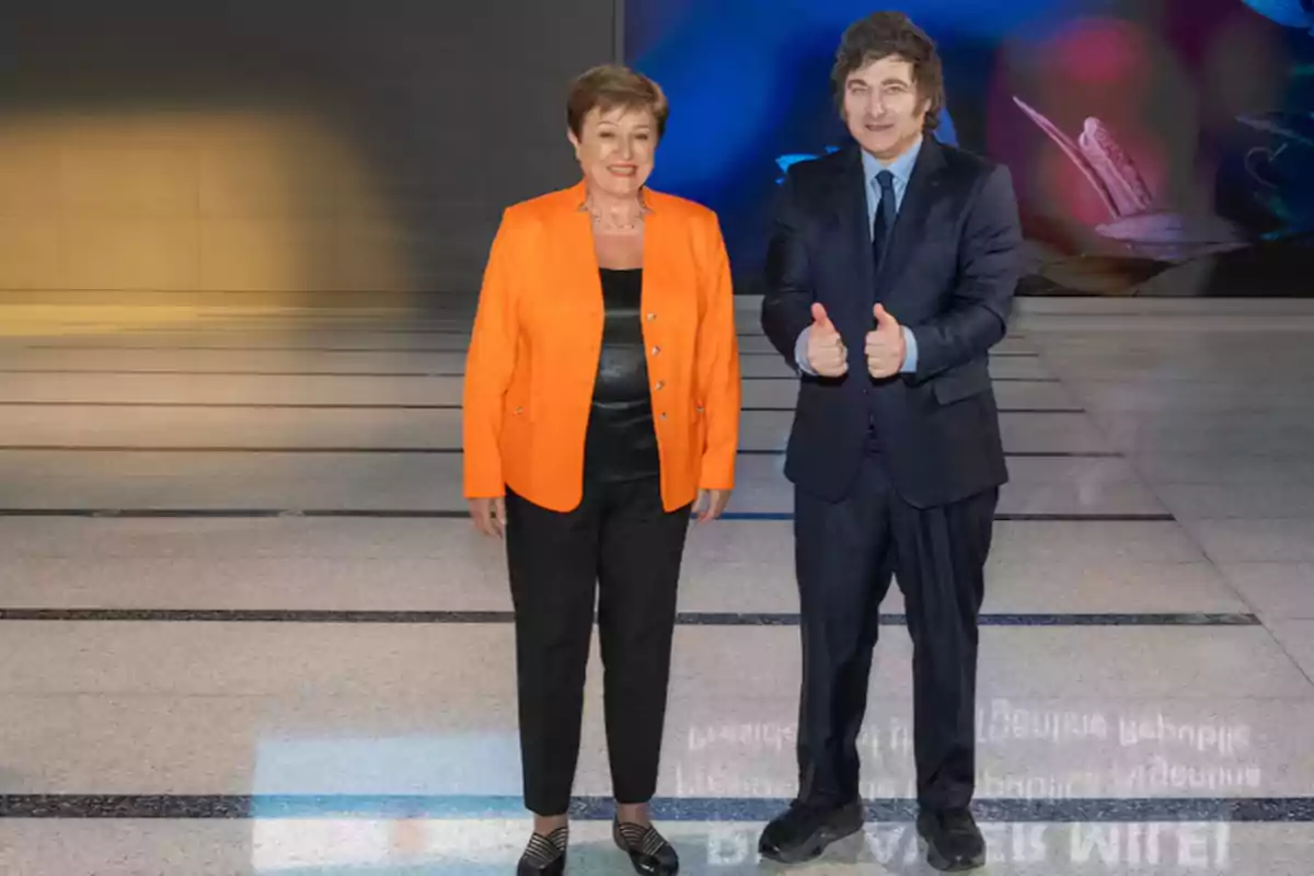 Dos personas posando en un espacio interior una de ellas lleva un traje naranja y la otra un traje oscuro haciendo un gesto de aprobación con los pulgares arriba.