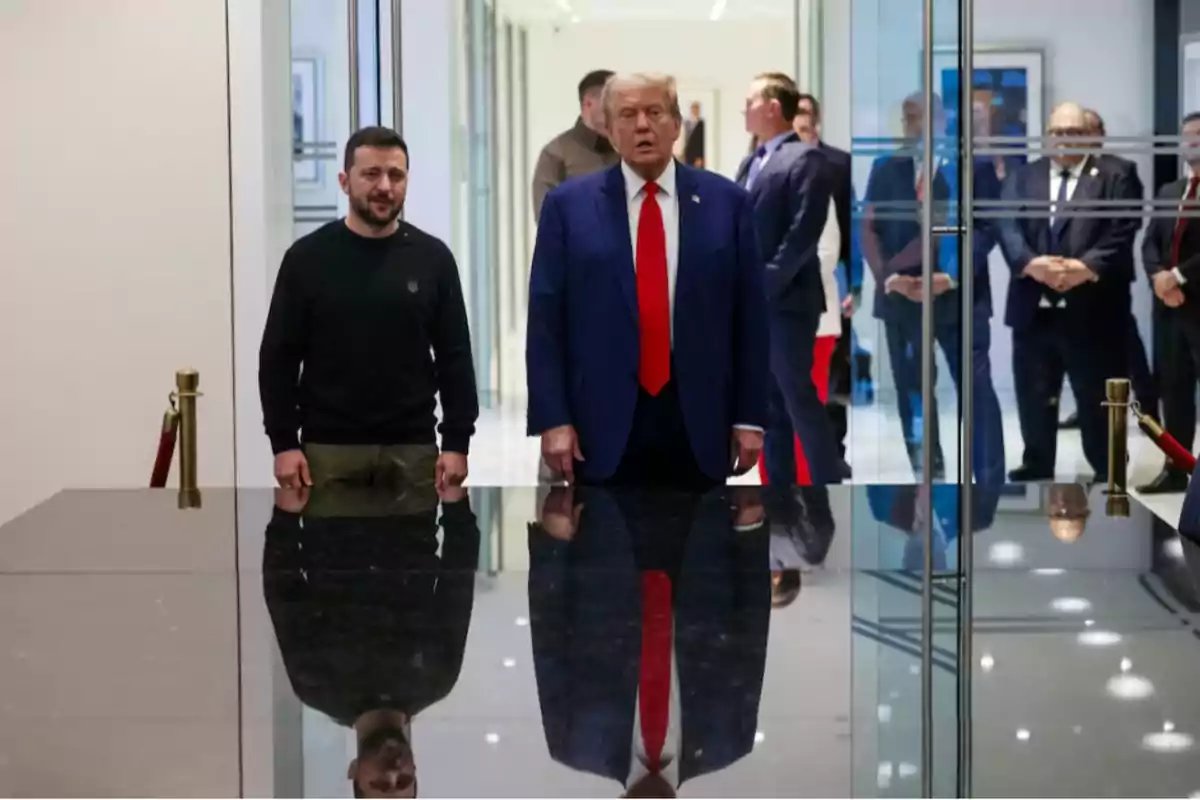 Dos hombres de pie en un entorno moderno con un reflejo en una superficie brillante, rodeados de otras personas en el fondo.