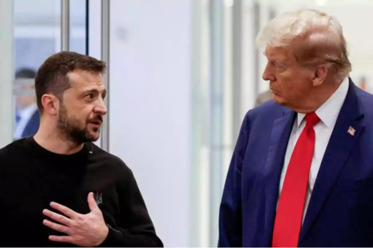 Dos hombres conversando en un entorno interior uno de ellos lleva un traje azul y corbata roja mientras que el otro viste una camiseta negra.