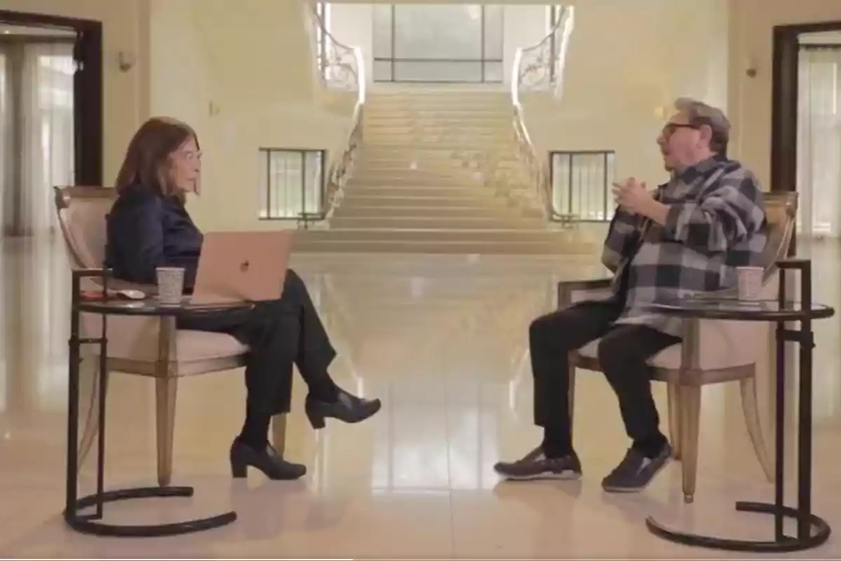 Two people sitting face to face in a large room with a staircase in the background, one of them has a laptop on a small table.