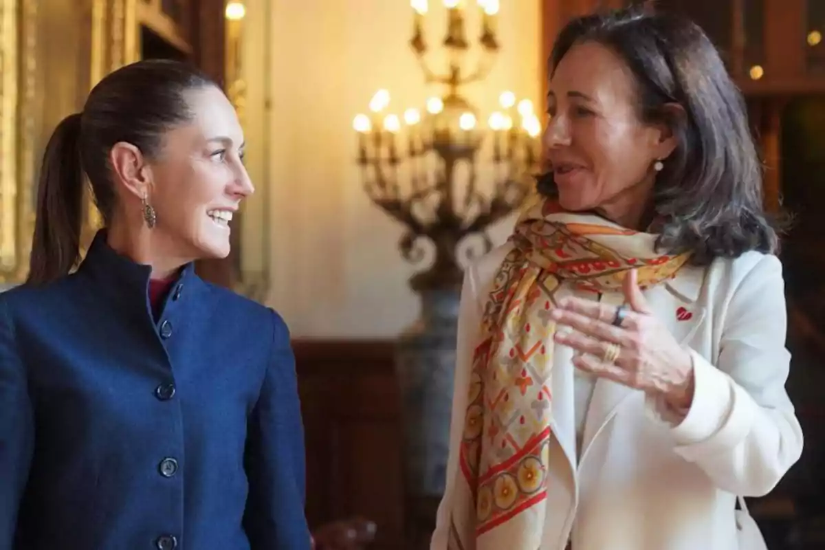 Two women chatting and smiling in an elegant setting with chandeliers in the background.