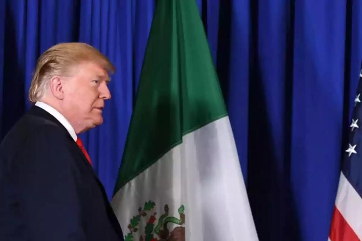 Un hombre de perfil frente a las banderas de México y Estados Unidos con un fondo azul.