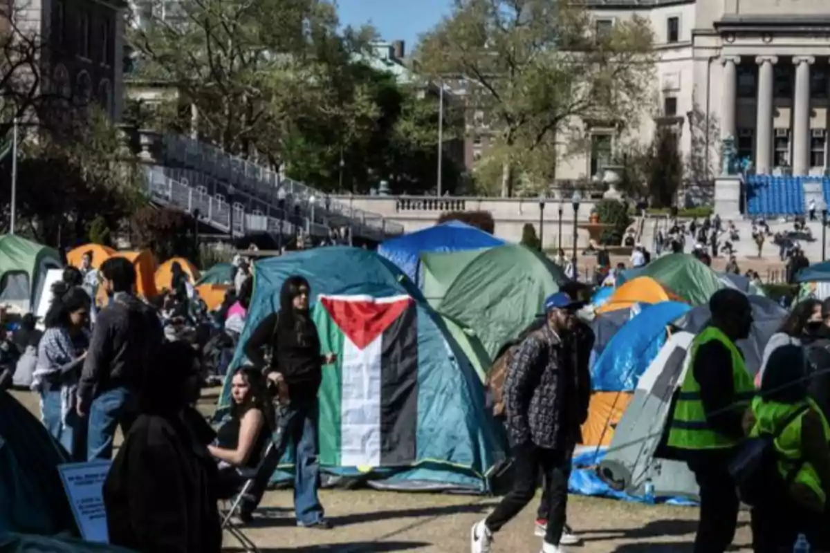 Un grupo de personas se encuentra reunido en un campamento de tiendas de campaña en un área al aire libre, con una bandera visible en una de las tiendas y un edificio de fondo.