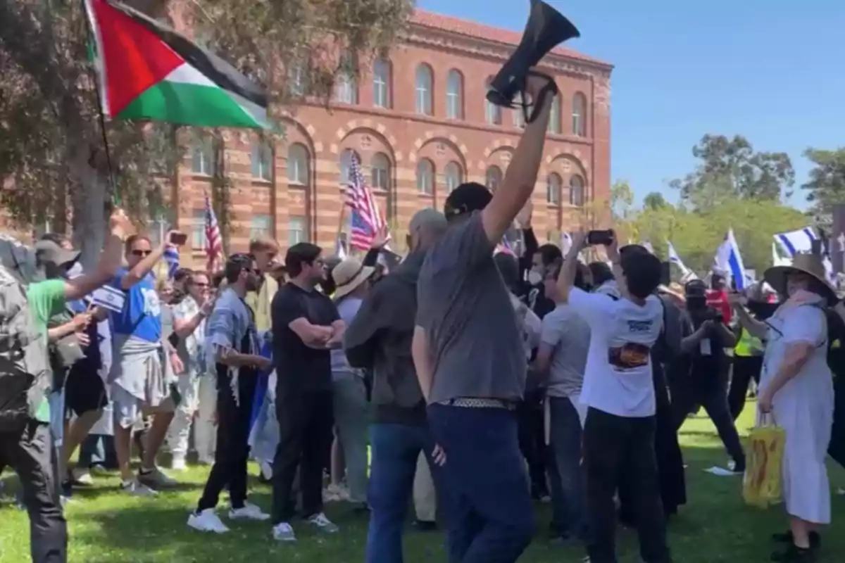 Un grupo de personas se reúne al aire libre, algunas sosteniendo banderas y un megáfono, frente a un edificio de ladrillo.