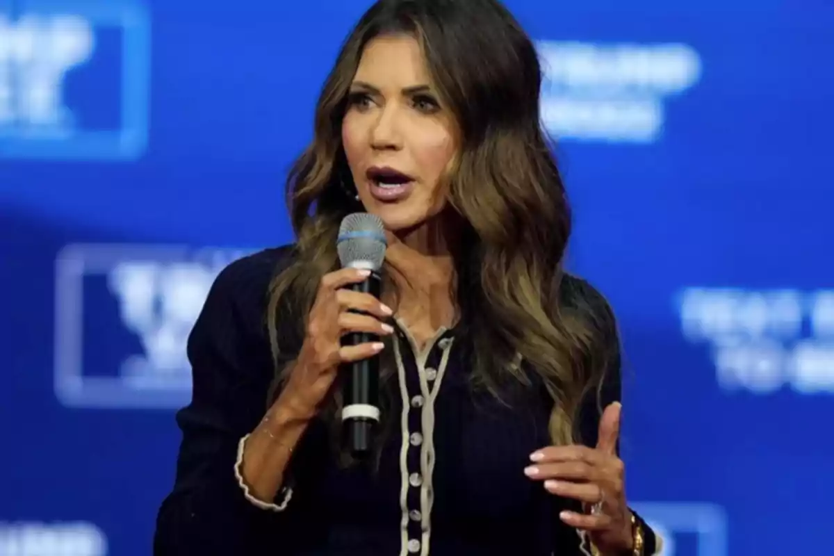 Una mujer hablando en un evento, sosteniendo un micrófono, con un fondo azul.