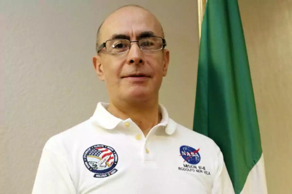 Un hombre con gafas y camiseta blanca con logotipos de la NASA, de pie junto a una bandera verde.