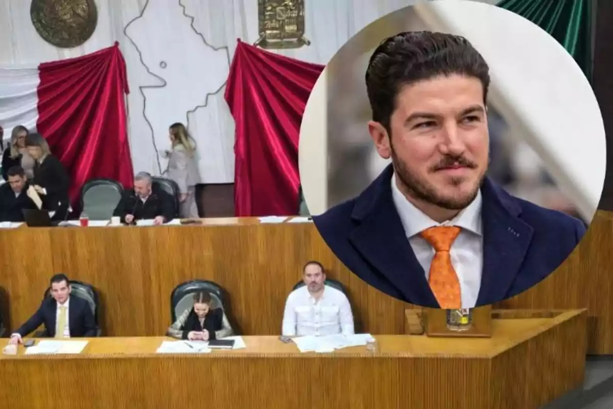 Una sala de reuniones con varias personas sentadas en un estrado de madera, con una bandera de fondo y un escudo en la pared; en la esquina superior derecha hay un recuadro con la imagen de un hombre con traje y corbata naranja.