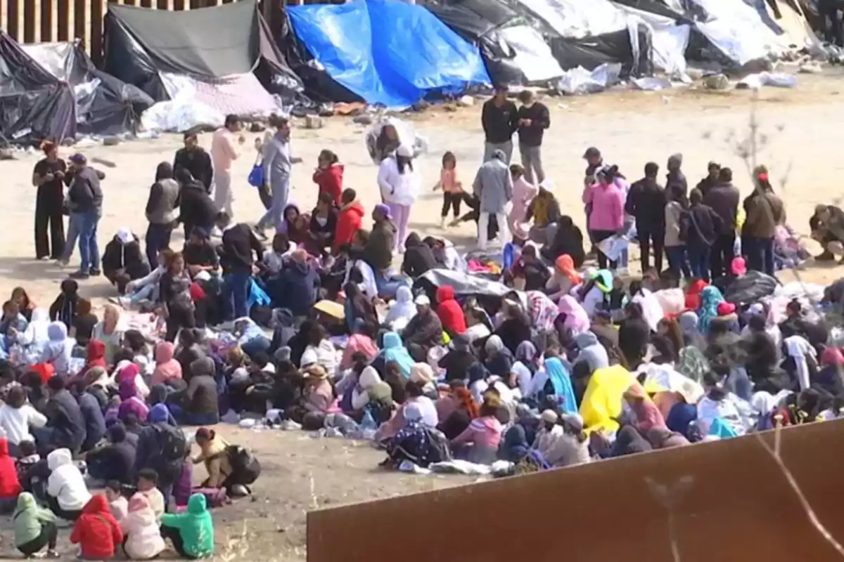 Un grupo numeroso de personas se encuentra reunido al aire libre, algunas están sentadas en el suelo mientras otras permanecen de pie, y al fondo se observan estructuras cubiertas con lonas de colores.