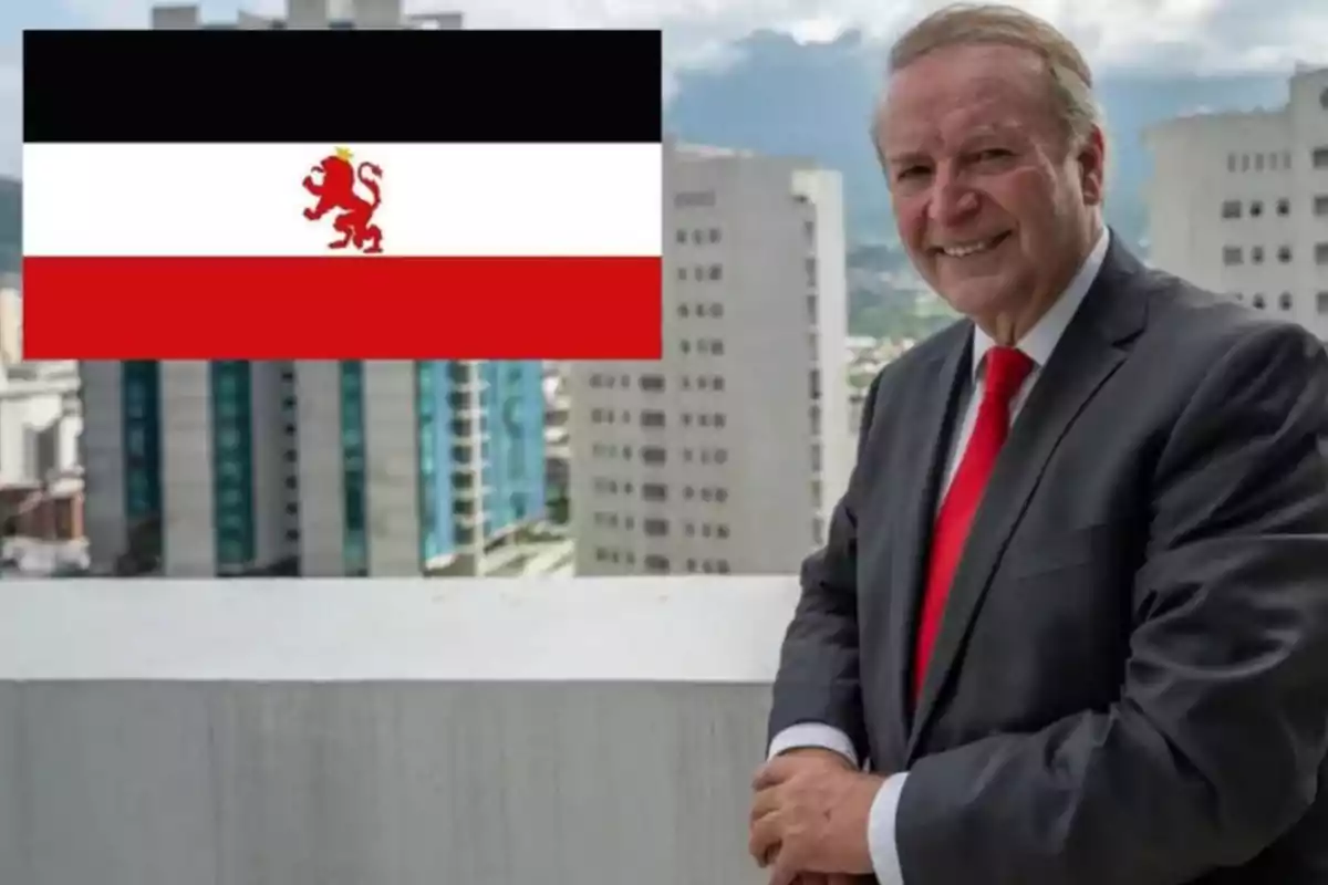 Un hombre de traje y corbata roja sonríe en una terraza con edificios de fondo y una bandera con un león en el lado izquierdo.