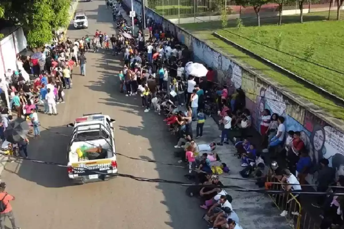 Una multitud de personas hace fila a lo largo de una calle, mientras una camioneta de policía está estacionada cerca.