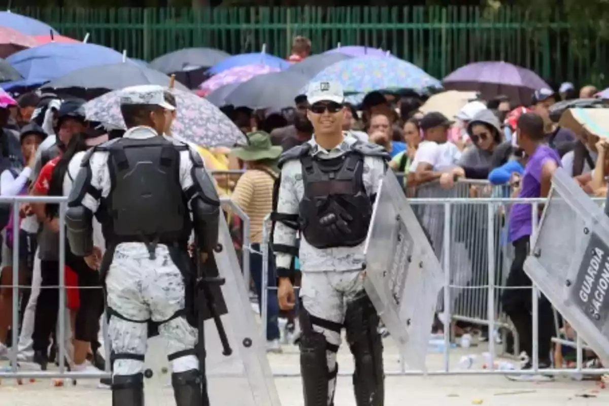 Dos guardias con equipo antidisturbios están de pie frente a una multitud de personas que sostienen paraguas de colores variados.