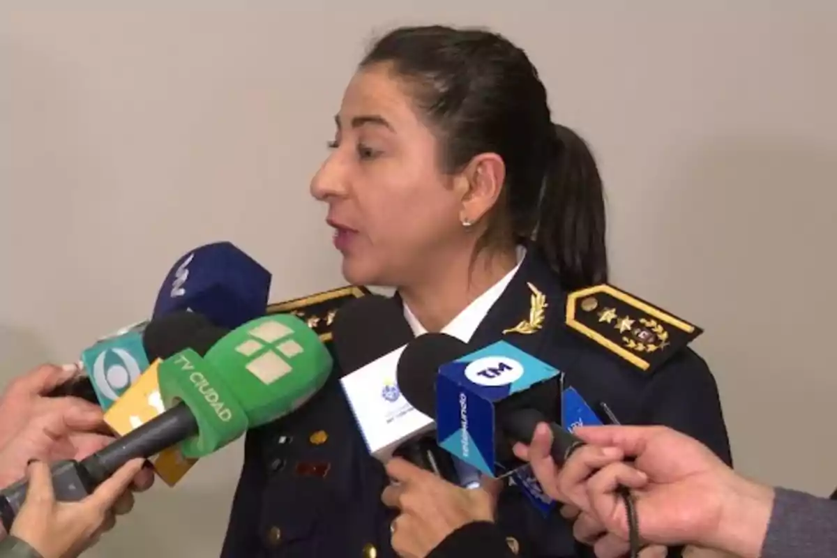 Una mujer en uniforme militar es entrevistada por varios medios de comunicación, rodeada de micrófonos de diferentes canales de televisión.