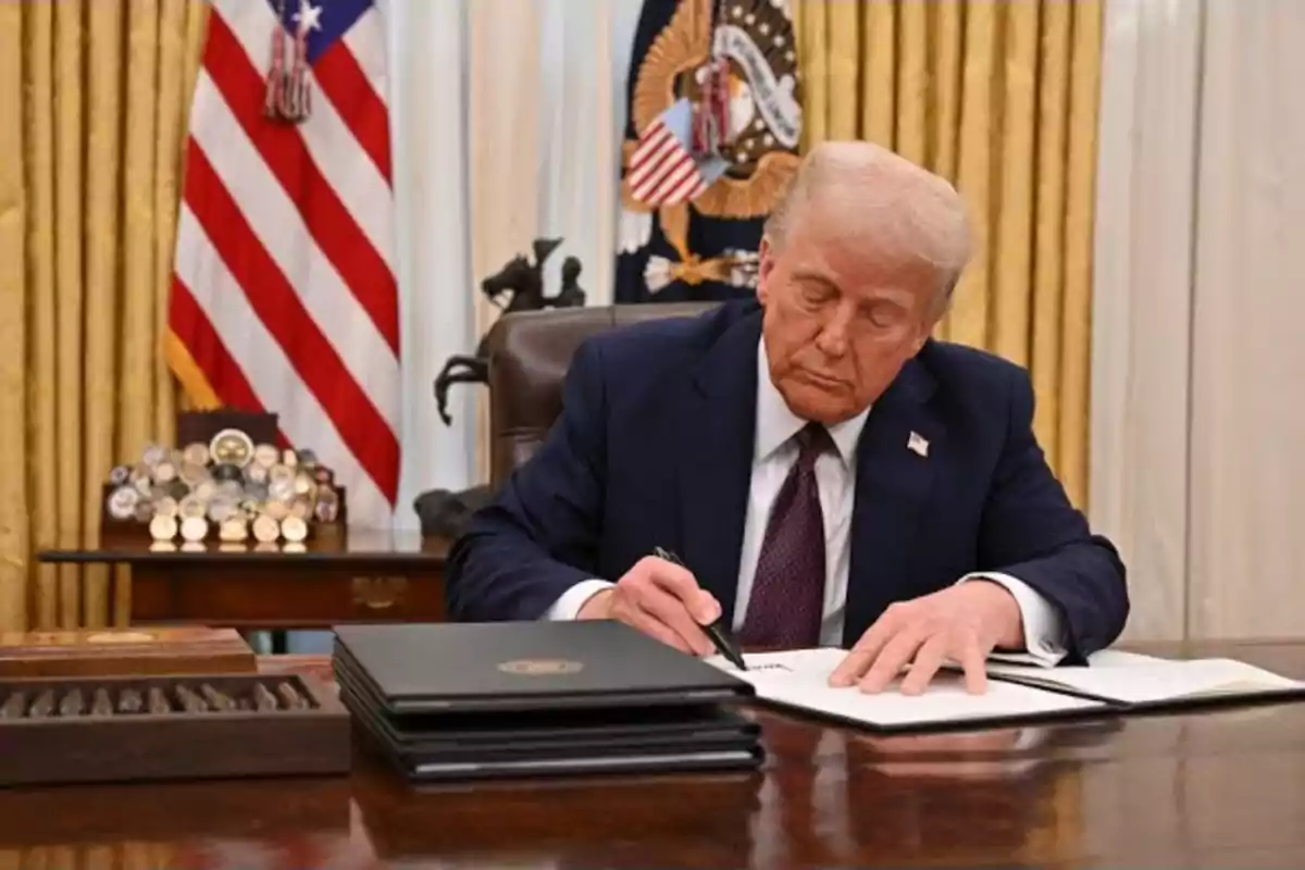 Un hombre de traje firmando documentos en un escritorio con una bandera estadounidense y un emblema presidencial en el fondo.