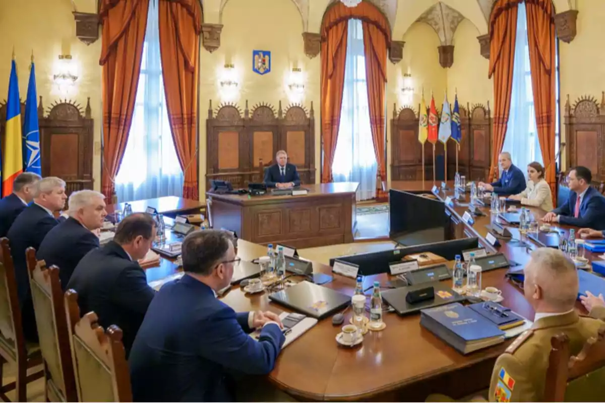 Un grupo de personas en una reunión formal en una sala con decoración elegante y banderas en el fondo.