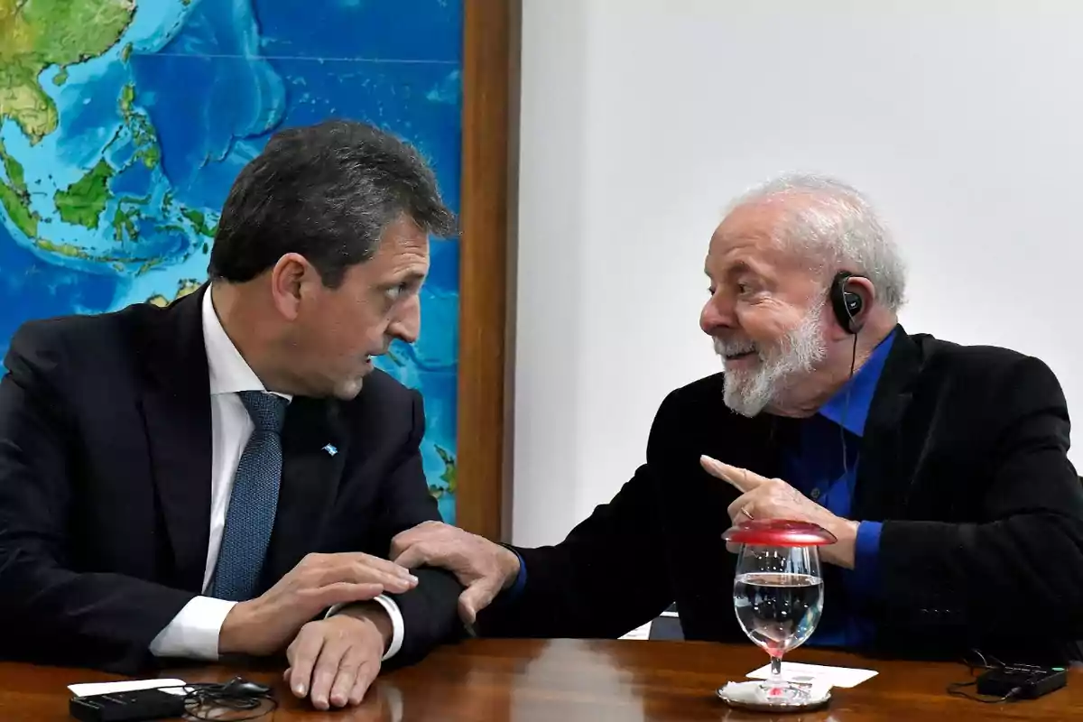 Dos hombres conversan animadamente en una mesa con un mapa de fondo.