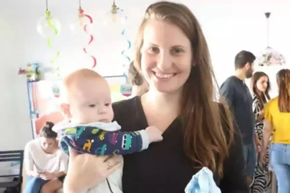 Una mujer sonriente sostiene a un bebé en un ambiente festivo con decoraciones coloridas y personas conversando al fondo.