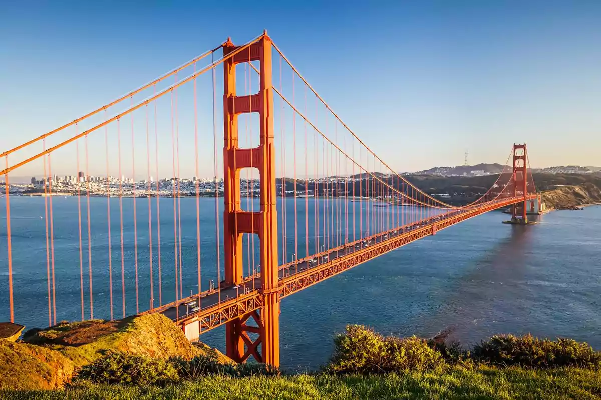 Puente colgante rojo sobre un cuerpo de agua con una ciudad al fondo en un día soleado.