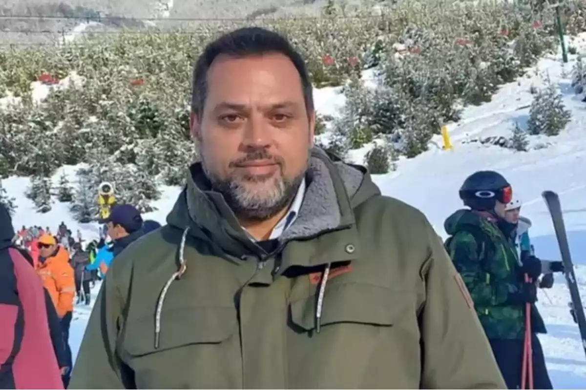 Un hombre con chaqueta verde en una estación de esquí nevada con personas y árboles al fondo.