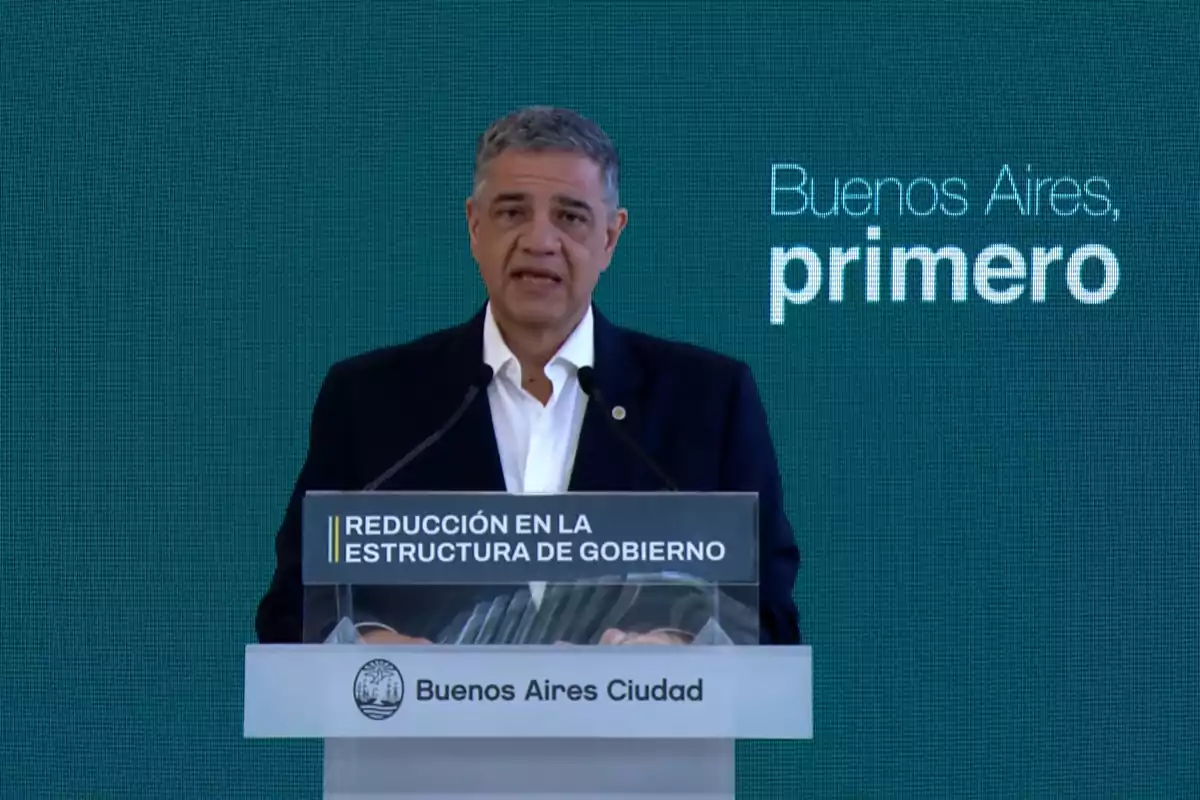 Un hombre hablando en un podio con el texto "Reducción en la estructura de gobierno" y "Buenos Aires, primero" en el fondo.