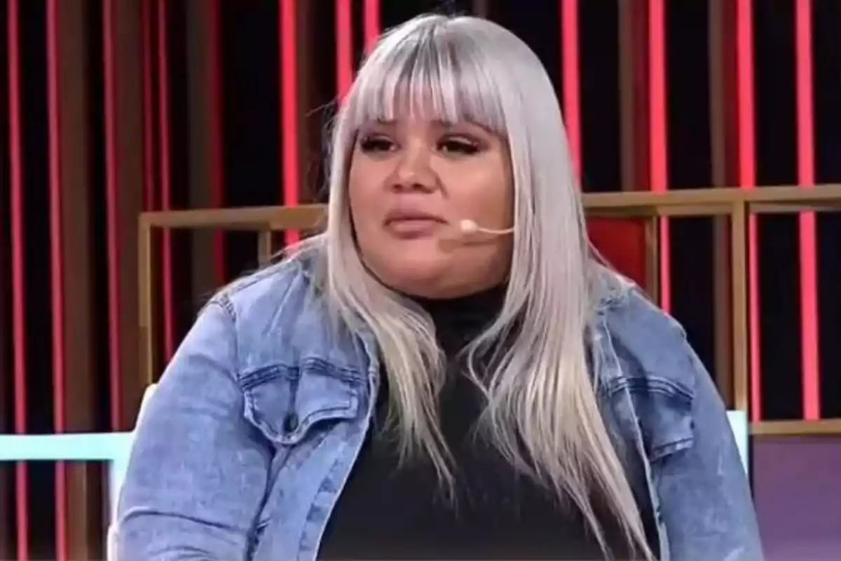 Una persona con cabello largo y rubio platino usando una chaqueta de mezclilla y un micrófono de diadema en un set de televisión.