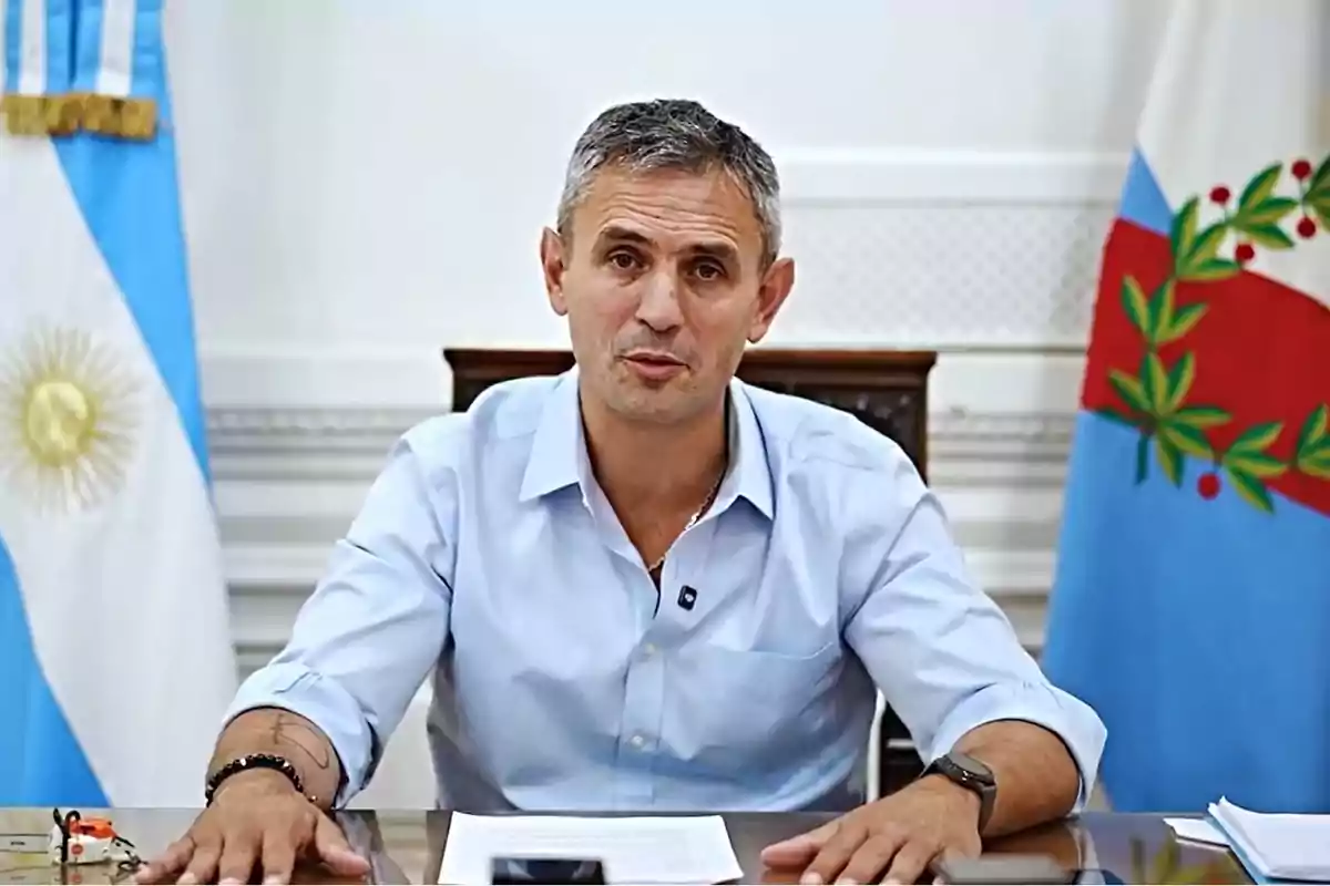 Un hombre de cabello corto y canoso, vestido con una camisa azul claro, está sentado en un escritorio con banderas argentinas detrás de él.