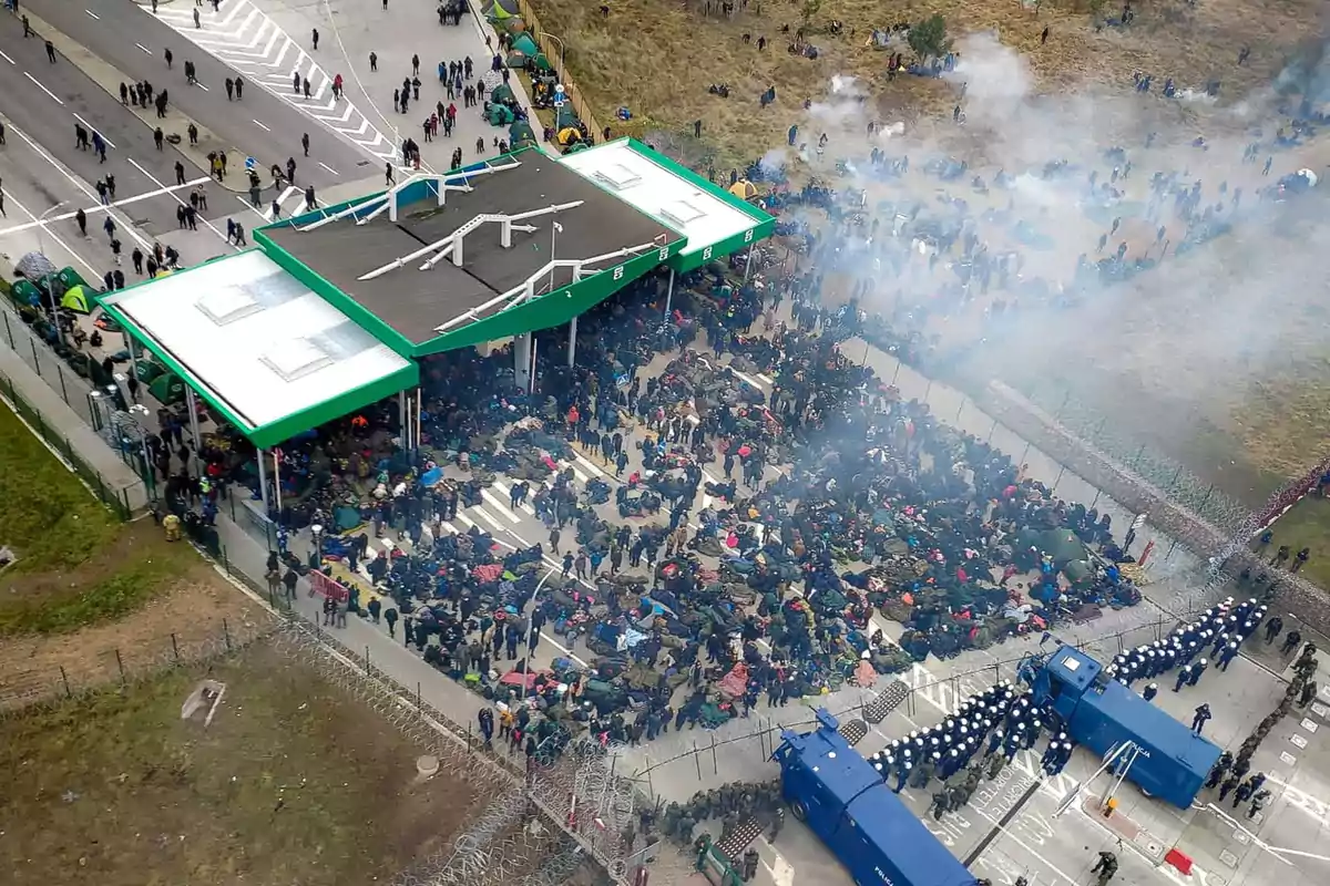 Una multitud se congrega en un puesto fronterizo, con humo disperso en el aire y vehículos de seguridad presentes.