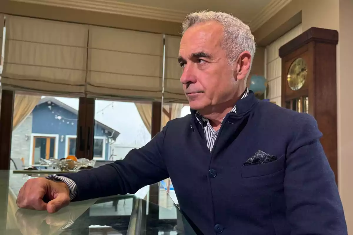 A gray-haired man in a dark jacket is sitting at a glass table in an elegant interior with a grandfather clock and a large window in the background.