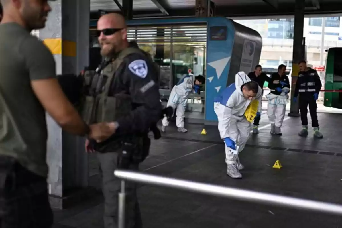 Un grupo de personas con trajes de protección y un oficial de seguridad están en una estación de transporte, realizando una investigación en la escena.