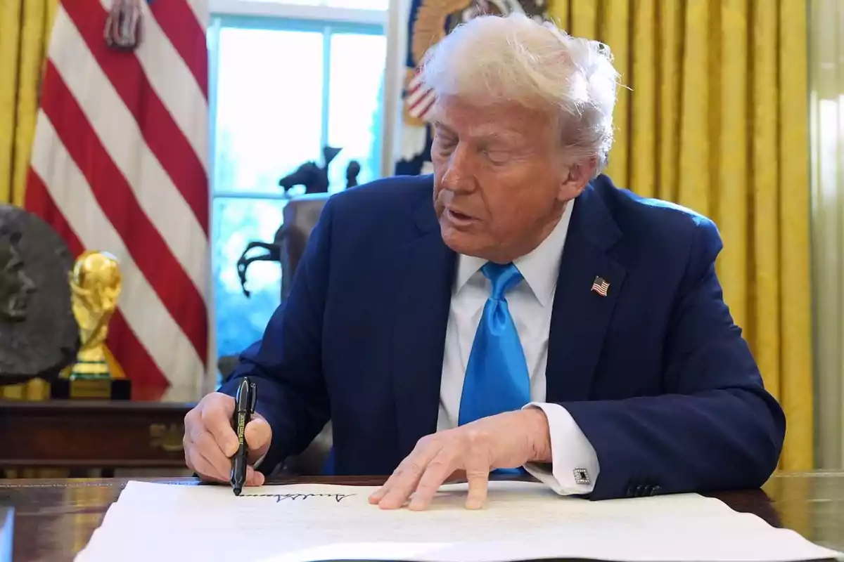 Un hombre de traje azul y corbata celeste está firmando un documento en un escritorio con una bandera de fondo.