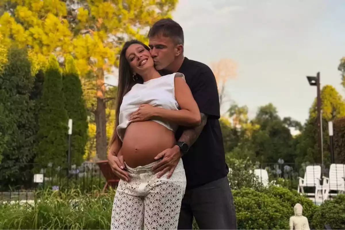 Una pareja en un jardín, donde el hombre abraza y besa a la mujer embarazada que sonríe.