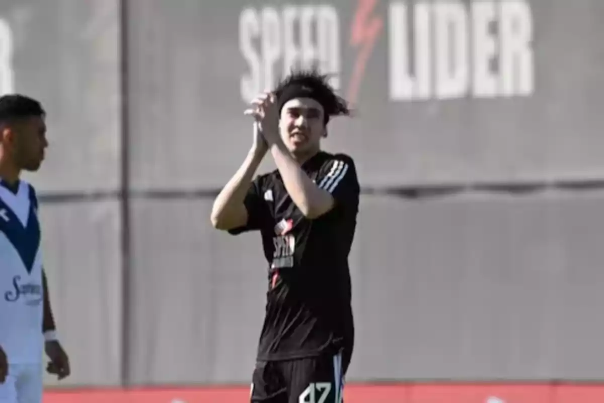 Un jugador de fútbol con uniforme negro aplaude en el campo mientras otro jugador con uniforme blanco y azul está al fondo.