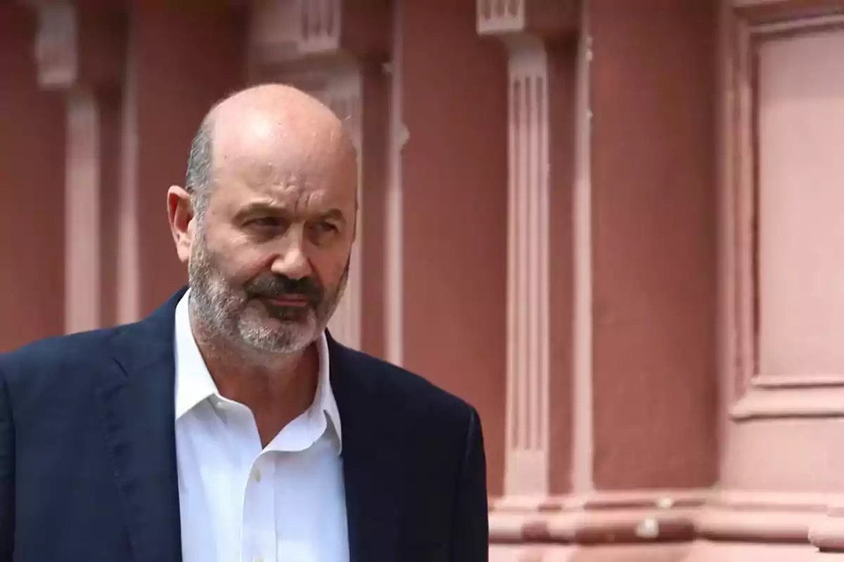 Un hombre con barba y traje oscuro está de pie frente a un edificio de color rosa.